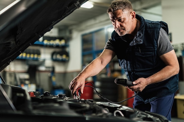 Mecánico adulto medio que usa una herramienta de diagnóstico mientras verifica el voltaje de una batería de automóvil en un taller de reparación de automóviles