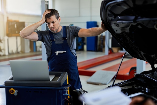 Foto gratuita mecánico angustiado que tiene problemas mientras repara el motor del automóvil y usa una computadora portátil en el taller de reparación de automóviles