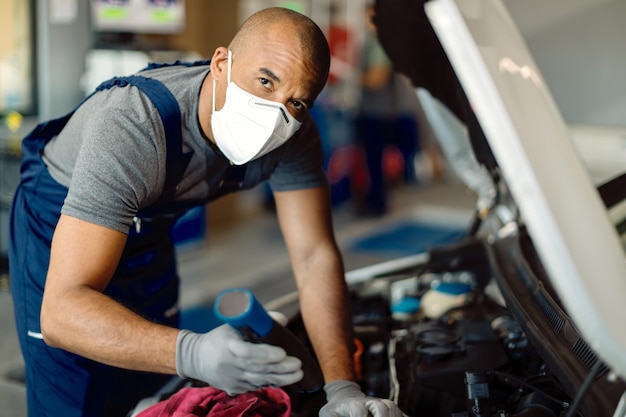 Mecánico de automóviles afroamericano con mascarilla examinando la avería del motor en un taller