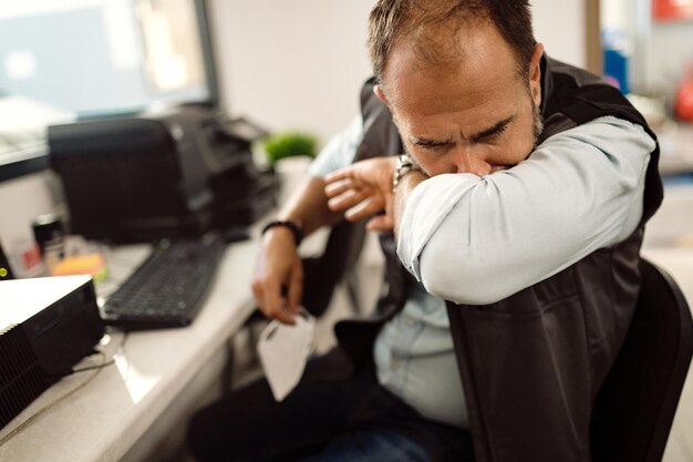 Mecánico de automóviles estornudando en el codo mientras trabajaba en la oficina de su taller