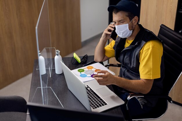 Mecánico de automóviles con mascarilla mientras habla por teléfono y trabaja en una computadora portátil en la oficina