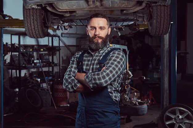 Mecánico experto barbudo vestido con uniforme, de pie con los brazos cruzados contra un coche en un ascensor en el garaje.