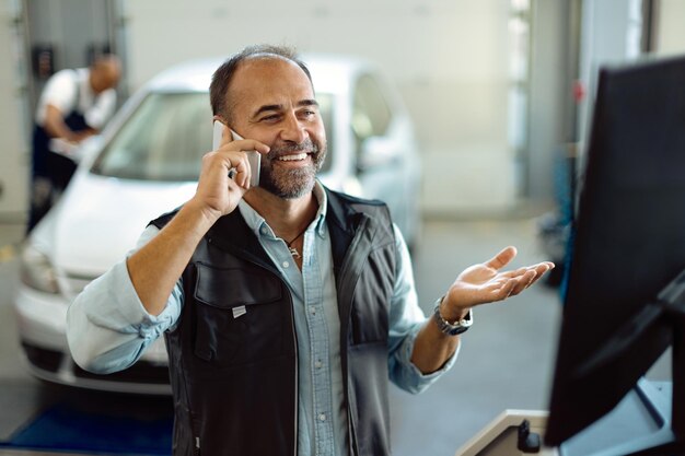 Mecánico feliz haciendo una llamada telefónica mientras trabaja en un taller de reparación de automóviles