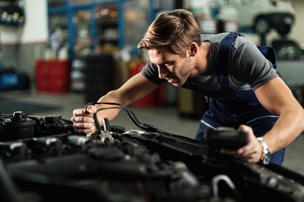 Mecánico joven que usa una herramienta de diagnóstico y conecta cables de puente en el motor del automóvil mientras trabaja en un taller de reparación de automóviles