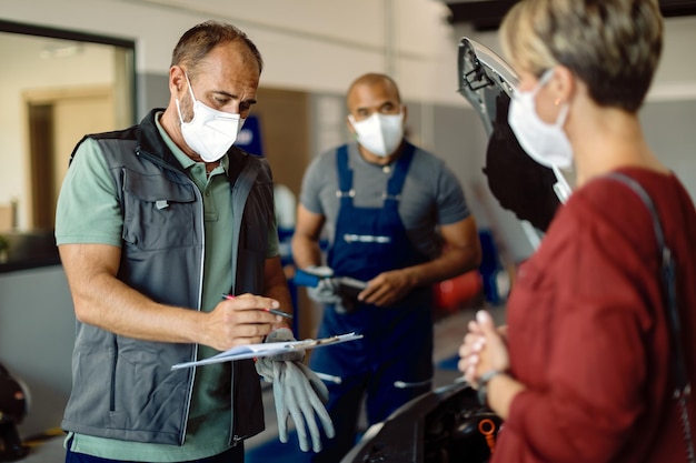 Mecánico con mascarilla revisando informes mientras habla con el cliente en el taller de reparación de automóviles