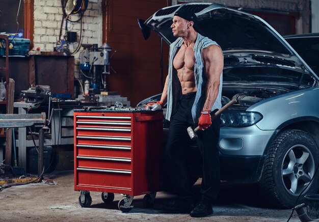 Mecánico musculoso vestido con una camisa de mezclilla posando cerca de un auto en un garaje.