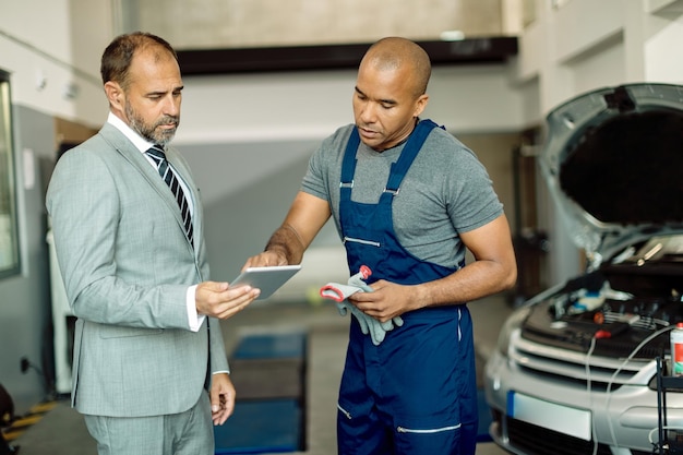 Mecánico negro y su gerente trabajando en el panel táctil en el taller de reparación de automóviles