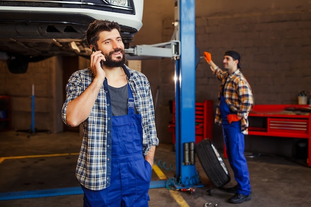 Mecánico sonriente hablando por teléfono inteligente con coche levantado y compañero de trabajo detrás