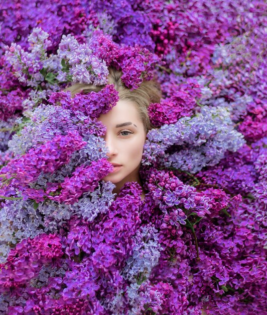 Media cara de joven rubia caucásica rodeada de un montón de lilas moradas y violetas, papel tapiz, melodía de primavera