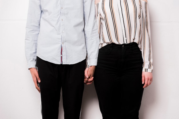 Mediados de sección de la joven pareja sosteniendo la mano de uno contra la pared blanca