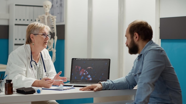 Médica femenina mirando la ilustración del coronavirus en una laptop con un paciente enfermo, hablando sobre medicación y prevención. Animación de virus en exhibición para curar enfermedades en el gabinete médico.