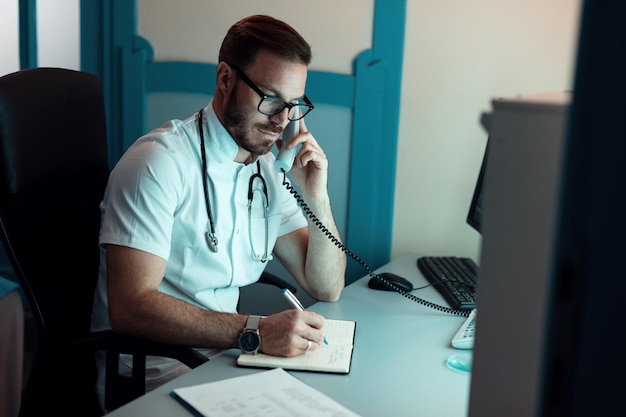 Médico adulto medio hablando con el paciente por teléfono y escribiendo notas en la clínica