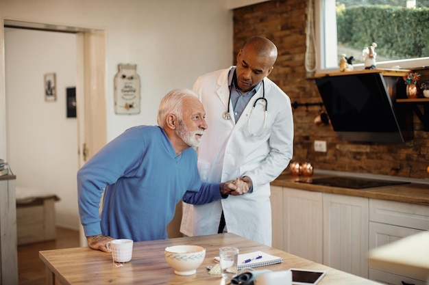 Médico afroamericano ayudando a un anciano a levantarse de la silla durante una visita a casa