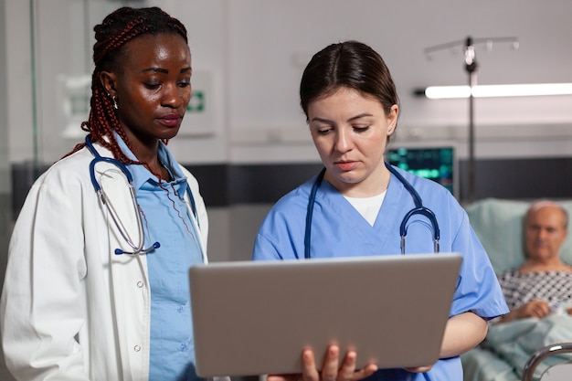 Médico afroamericano especialista en salud y asistente usando una computadora portátil, discutiendo hablar, tratamiento y diagnóstico, en la habitación del hospital con el paciente enfermo acostado en la cama con un goteo intravenoso adjunto.