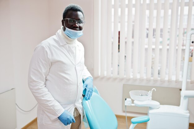 Médico afroamericano con gafas y máscara de pie cerca de la silla del dentista en la clínica dental