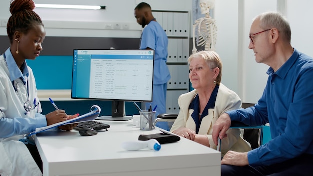 Médico afroamericano hablando con una anciana con discapacidad en la cita de visita de chekcup. Paciente mayor con discapacidad en silla de ruedas y esposo que recibe tratamiento médico por enfermedad.