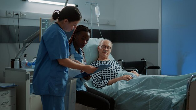 Médico afroamericano hablando con un paciente enfermo en la cama en la sala del hospital para tratar problemas de salud y lesiones. Anciano preguntando a médico y enfermera sobre cuidados intensivos y recuperación de enfermedades