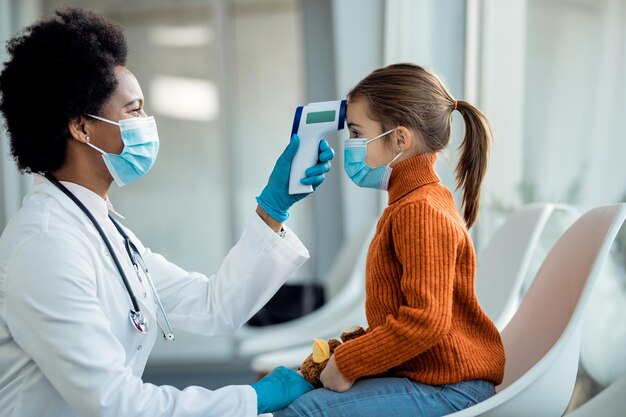 Médico afroamericano midiendo la temperatura de una niña pequeña en una sala de espera en la clínica