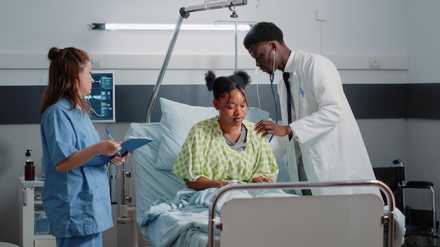 Médico afroamericano que usa estetoscopio en un paciente enfermo para controlar los latidos del corazón y hacer un examen para curar la enfermedad. Especialista en consulta de atención médica mientras la enfermera brinda asistencia