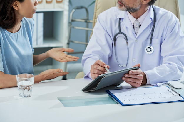 médico apuntando a la pantalla de la tableta digital mientras explica algo al paciente