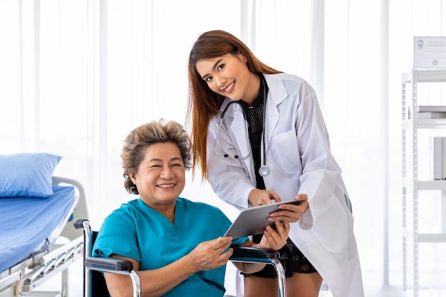 Un médico asiático cuida y explica a una anciana paciente en silla de ruedas con una tableta mirando a la cámara