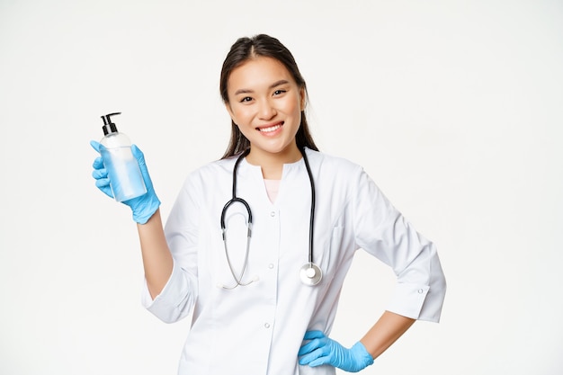 Médico asiático sonriente sosteniendo desinfectante de manos con guantes de goma, mostrando una botella con antiséptico para la prevención del coronavirus, fondo blanco.