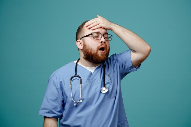 Médico barbudo uniformado con estetoscopio alrededor del cuello con gafas que parece confundido y decepcionado con la mano en la cabeza por error de pie sobre fondo azul