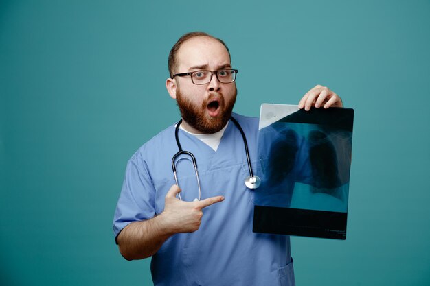 Médico barbudo uniformado con estetoscopio alrededor del cuello con gafas sosteniendo una radiografía pulmonar apuntando con el dedo índice mirando asombrado y sorprendido de pie sobre fondo azul