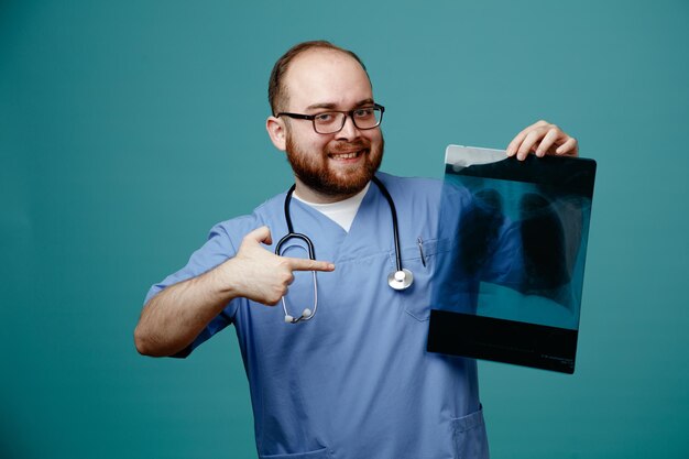 Médico barbudo uniformado con estetoscopio alrededor del cuello con gafas sosteniendo una radiografía pulmonar feliz y positiva señalando con el dedo índice sonriendo de pie sobre fondo azul