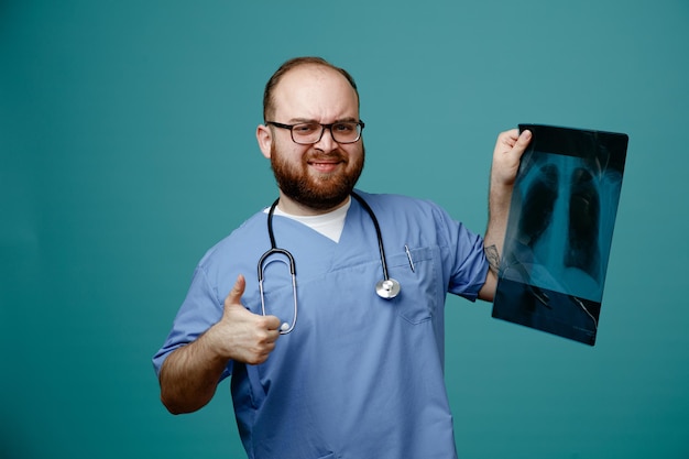 Foto gratuita médico barbudo uniformado con estetoscopio alrededor del cuello con gafas sosteniendo una radiografía pulmonar mirando a la cámara sonriendo confundido mostrando el pulgar hacia arriba de pie sobre fondo azul