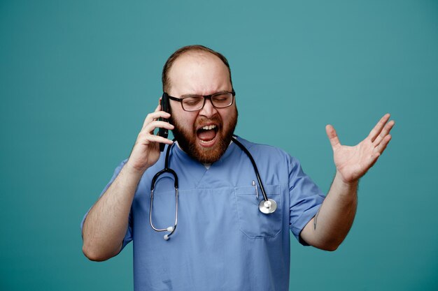 Médico barbudo en uniforme con estetoscopio alrededor del cuello con gafas enojado y frustrado mientras habla por teléfono móvil de pie sobre fondo azul.