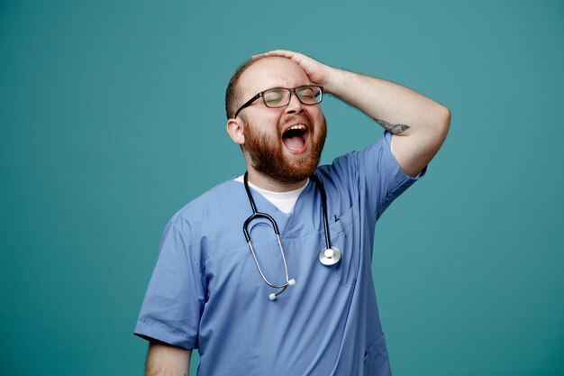 Médico barbudo en uniforme con estetoscopio alrededor del cuello con gafas feliz y emocionado sosteniendo la mano en su cabeza de pie sobre fondo azul
