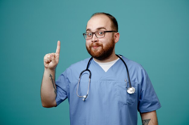 Médico barbudo en uniforme con estetoscopio alrededor del cuello con gafas mirando a la cámara feliz y positivo mostrando el dedo índice con buenas noticias de pie sobre fondo azul.