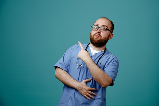 Médico barbudo en uniforme con estetoscopio alrededor del cuello con gafas que parece confiado apuntando con el dedo índice hacia el lado de pie sobre fondo azul