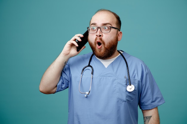 Médico barbudo en uniforme con estetoscopio alrededor del cuello con gafas que parece sorprendido mientras habla por teléfono móvil de pie sobre fondo azul.