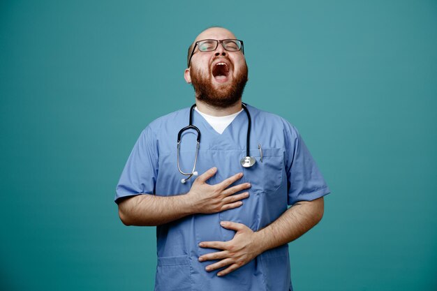 Médico barbudo en uniforme con estetoscopio alrededor del cuello con gafas riéndose cogidos de la mano en su vientre de pie sobre fondo azul.