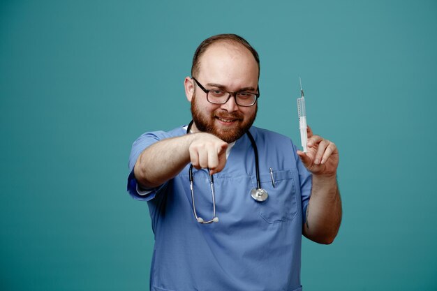 Médico barbudo en uniforme con estetoscopio alrededor del cuello con gafas sosteniendo una jeringa apuntando con el dedo índice a la cámara sonriendo astutamente feliz y positivo de pie sobre fondo azul