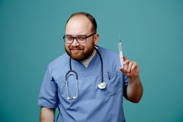 Médico barbudo en uniforme con estetoscopio alrededor del cuello con gafas sosteniendo una jeringa mirando a la cámara con una gran sonrisa en la cara de pie sobre fondo azul.