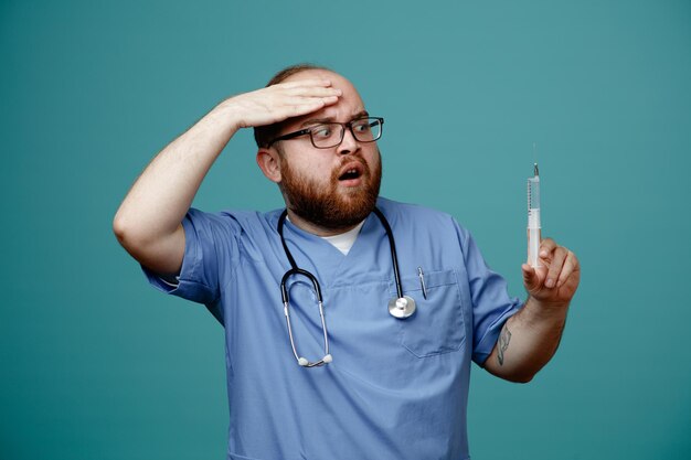 Médico barbudo en uniforme con estetoscopio alrededor del cuello con gafas sosteniendo una jeringa mirándolo preocupado y confundido de pie sobre fondo azul