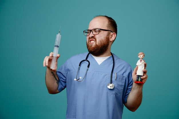 Médico barbudo en uniforme con estetoscopio alrededor del cuello con gafas sosteniendo una jeringa y una muñeca que parece confundida y disgustada de pie sobre fondo azul