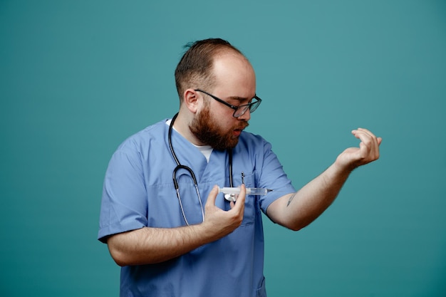 Foto gratuita médico barbudo en uniforme con estetoscopio alrededor del cuello con gafas sosteniendo una jeringa que se va a inyectar de pie sobre fondo azul