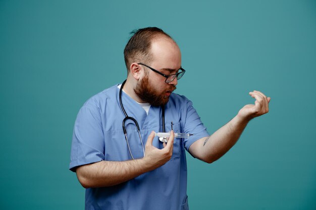 Médico barbudo en uniforme con estetoscopio alrededor del cuello con gafas sosteniendo una jeringa que se va a inyectar de pie sobre fondo azul