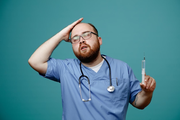 Médico barbudo en uniforme con estetoscopio alrededor del cuello con gafas sosteniendo una jeringa que parece confundido y preocupado sosteniendo la mano en su cabeza por error de pie sobre fondo azul