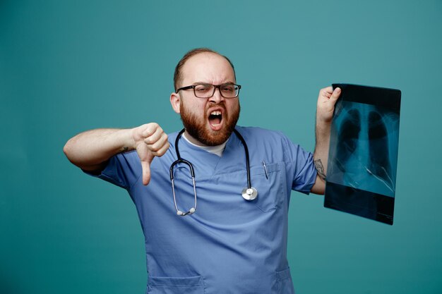 Médico barbudo en uniforme con estetoscopio alrededor del cuello con gafas sosteniendo rayos X de pulmón gritando con expresión agresiva mostrando el pulgar hacia abajo de pie sobre fondo azul