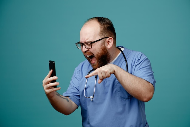 Médico barbudo en uniforme con estetoscopio alrededor del cuello con gafas sosteniendo un teléfono inteligente gritando con expresión agresiva de pie sobre fondo azul.