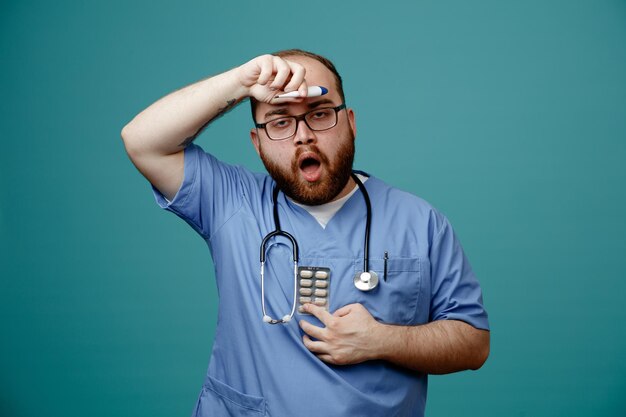 Médico barbudo en uniforme con estetoscopio alrededor del cuello con gafas con termómetro y pastillas que parecen confundidos y preocupados de pie sobre fondo azul.