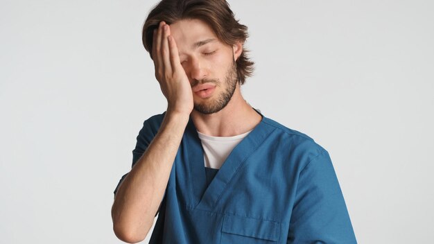 Médico cansado con la mano en la cara sintiéndose somnoliento después de un duro día en el hospital Joven trabajador médico vestido con uniforme que parece cansado sobre fondo blanco