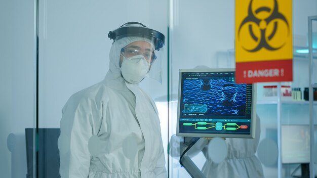 Médico cansado en traje de protección contra el covid-19 mirando agotado a la cámara detrás de la pared de vidrio trabajando en la zona de peligro. Científico que examina la evolución del virus utilizando alta tecnología para la investigación científica