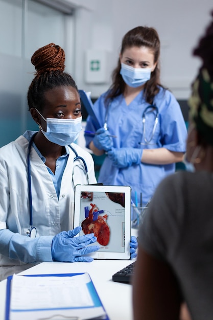 Médico cardiólogo afroamericano con tableta que muestra radiografía de corazón al paciente enfermo explicando el tratamiento de medicaton durante la cita clínica en la oficina del hospital. Personas con cara médica ma