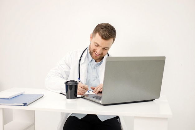 Foto gratuita médico caucásico sentado en el lugar de trabajo y usando una computadora portátil